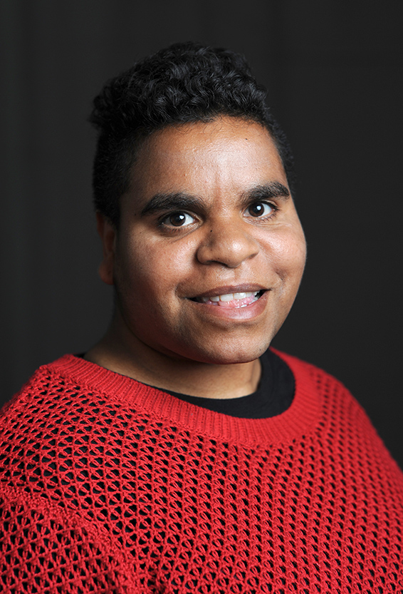A portrait photograph of an Aboriginal woman with short hair look directly at the camera and smiles. She is wearing a black T-shirt and red jumper.