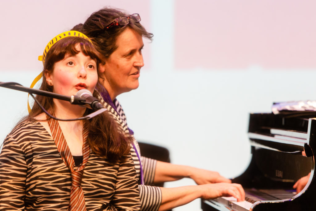 A photo of a young girl singing into a microphone as a woman plays piano in the background