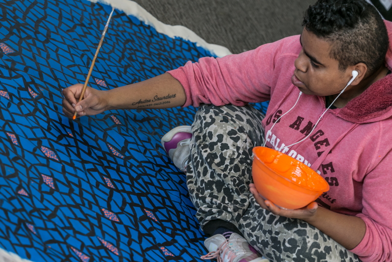 A photo of a woman sitting and holding a bowl of paint as she paints a large canvas. She is wearing earphones.