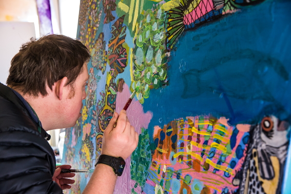 A photo of a man standing and painting a large multicoloured artwork