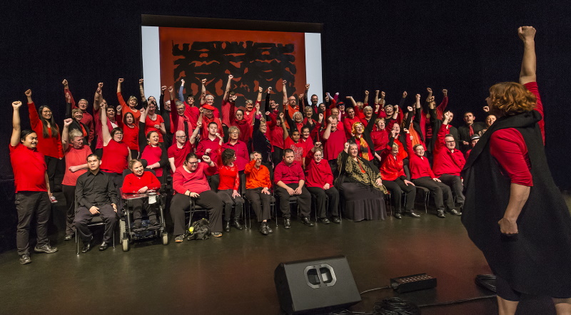 A photo of a choir and conductor, dressed in red and black, pumping their fists in the air