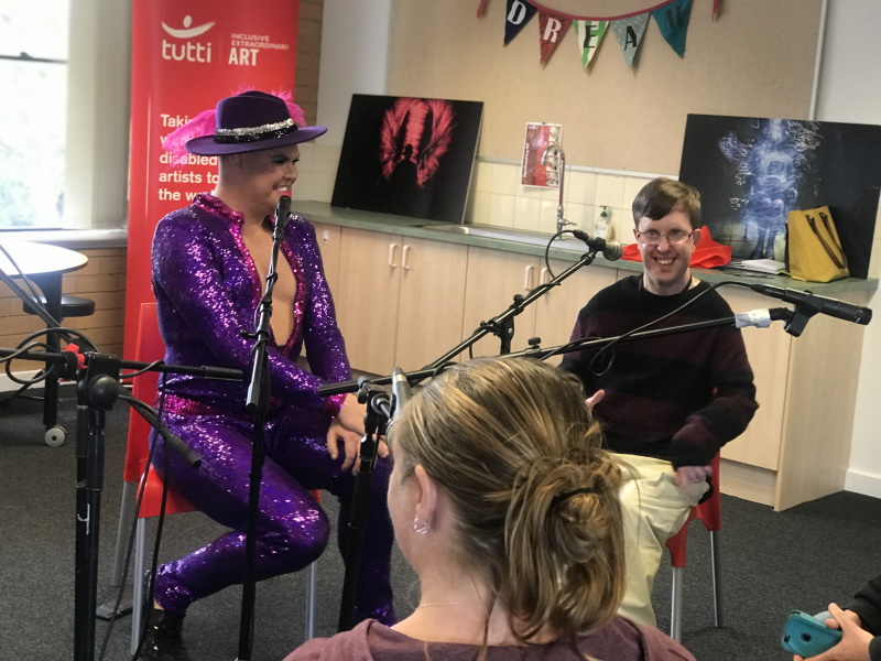 A photo of international superstar Hans, in a sequined jumpsuit and hat, sitting with others behind microphones