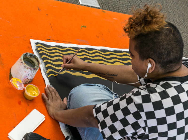 A photo looking over the shoulder of an Aboriginal woman with short hair as she sits and paints on a canvas