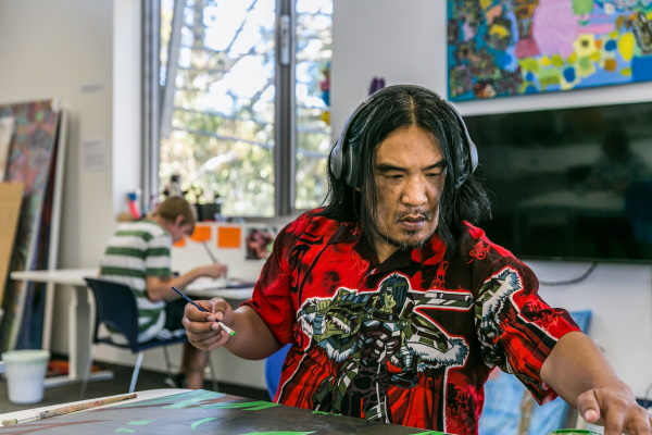 A photo of a long-haired, bearded man wearing headphones holding a paint brush and reaching for a pot of paint as another artist sits at work behind him