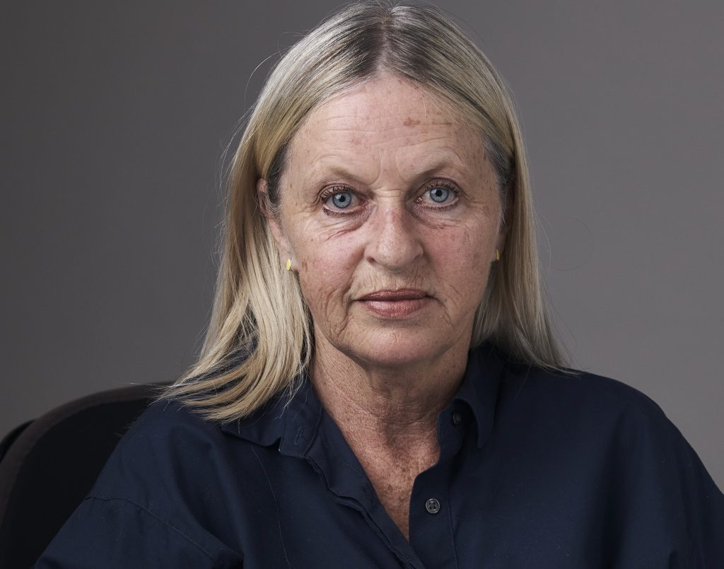 A photo of a woman with long ash-coloured hair and blue eyes who is sitting in a black electric wheelchair with her hands clasped.