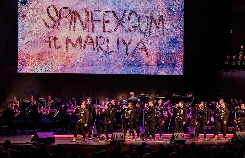 An orchestra on stage underneath a sign saying "Spinifex Gum ft Marliya"